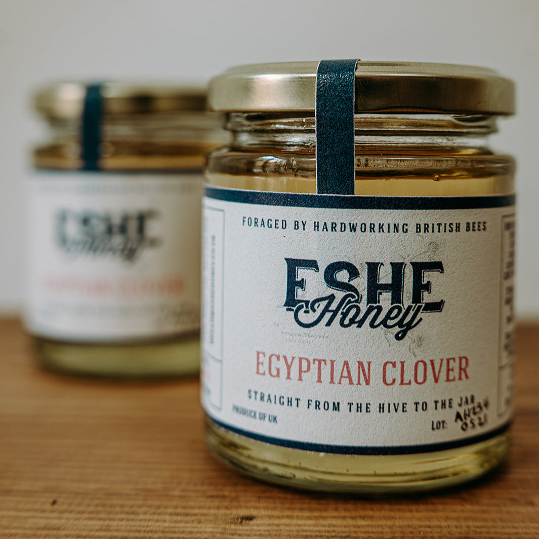 Two jars of Eshe Honey Egyptian Clover on a wooden surface with a white background