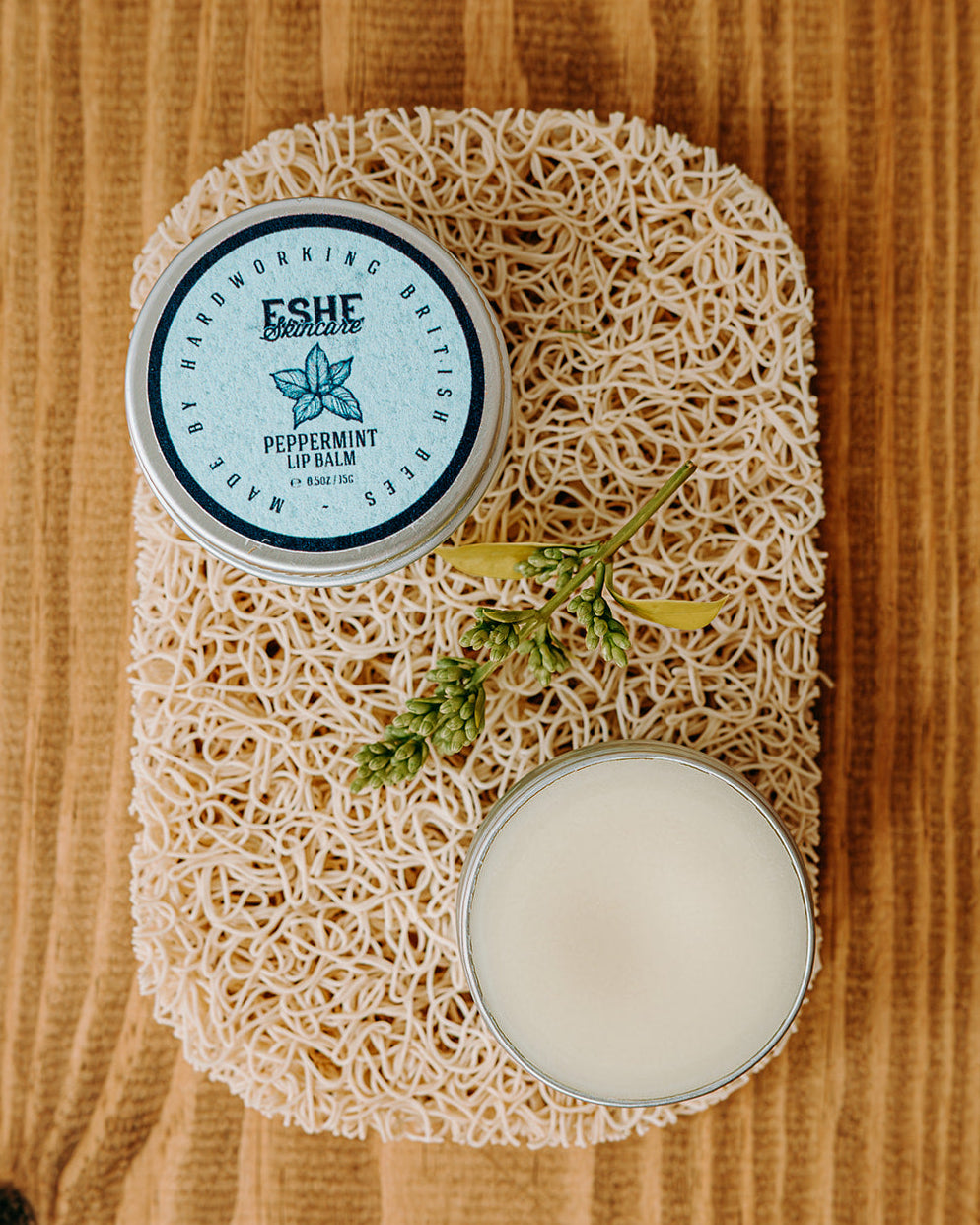 Two containers of Eshe peppermint lip balm on a wooden surface with a sponge.