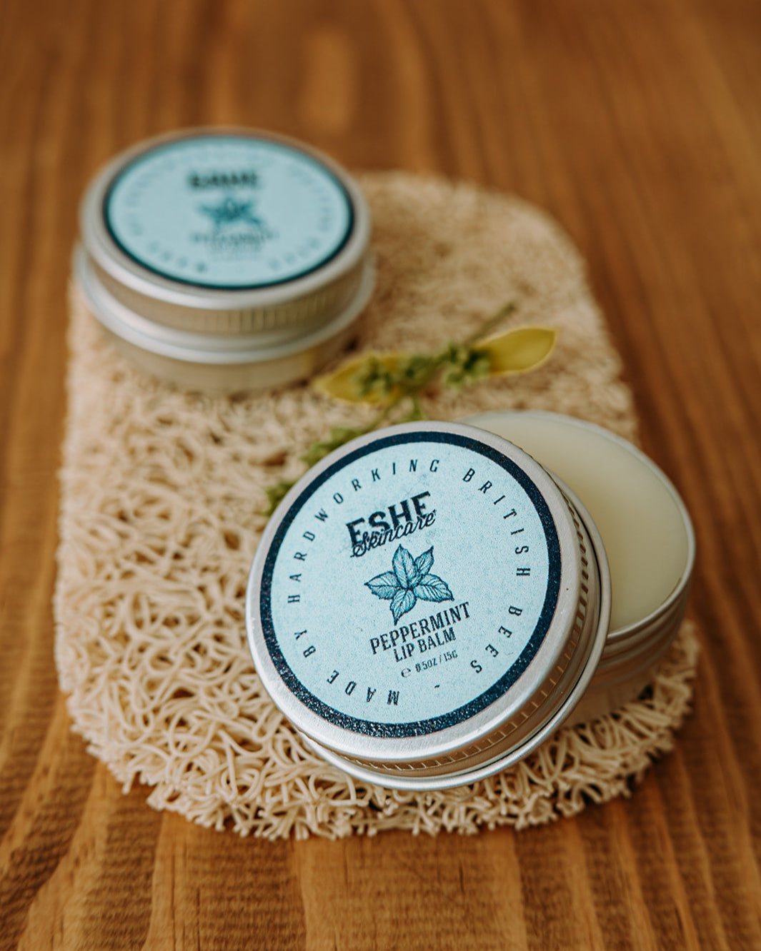 Two tins of Eshe peppermint lip balm on a wooden surface with a textured mat underneath.