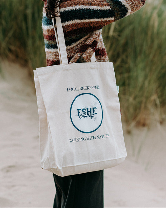 Person holding a tote bag with Eshe brand logo on a beach
